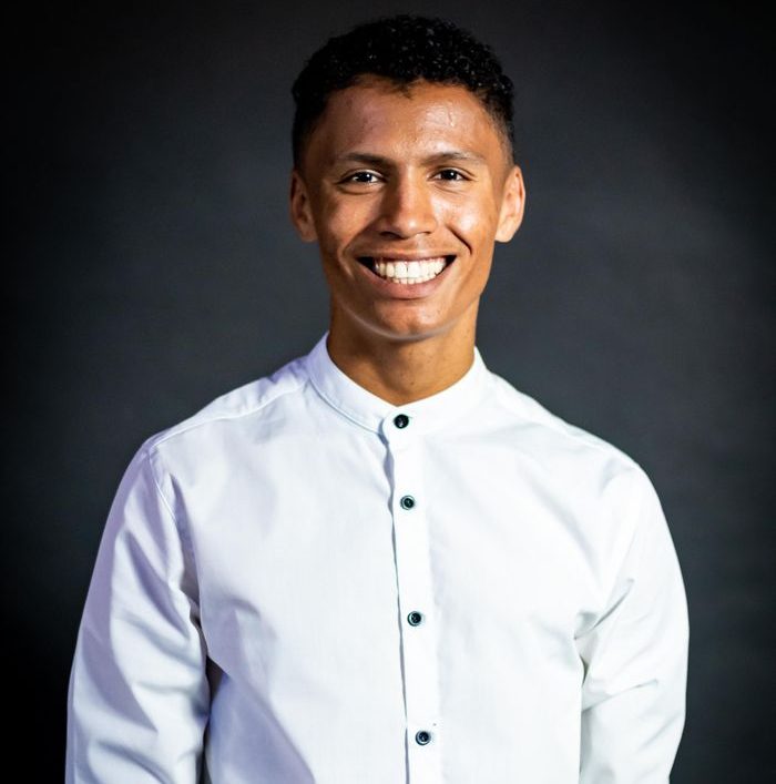 Bright studio portrait of a young man smiling in a white shirt against a dark background.