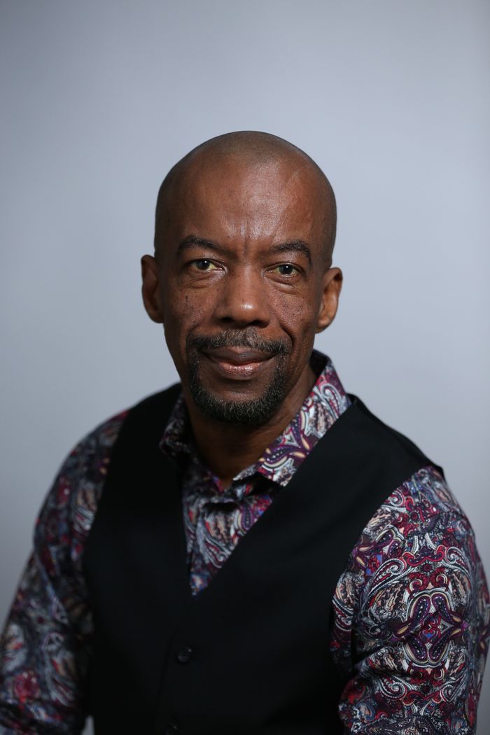 Studio portrait of an adult black man with a confident expression, wearing a patterned shirt and vest.